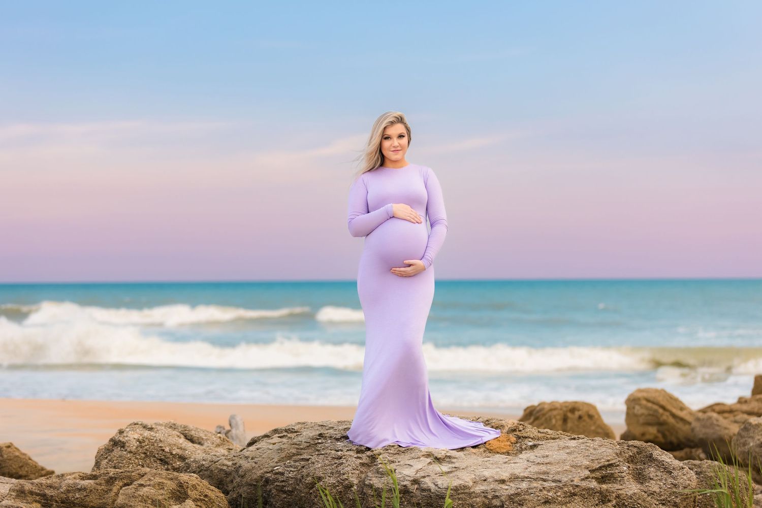 Expectant mother in flowing lavender dress poses on rocky beach during sunset photoshoot.
