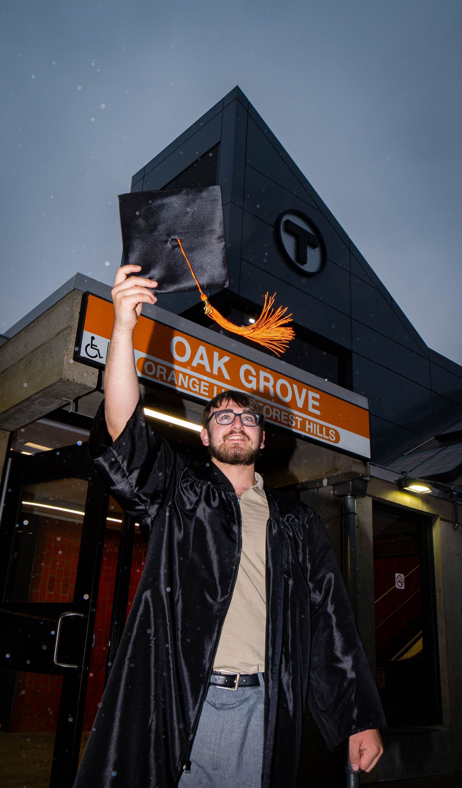 A person in a graduation cap and gown stands at Oak Grove station at dusk with arms raised in celebration.