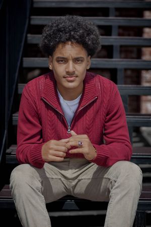 A young person in a burgundy sweater sits on outdoor steps while holding a phone, showing a relaxed casual pose.