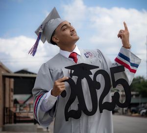 Graduate in white cap and gown holds up 2025 numbers while pointing to the sky against blue clouds.