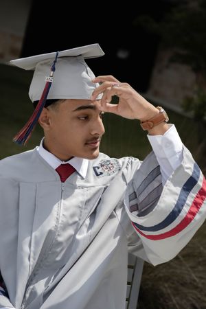 A graduate in a white cap and gown with red and blue stripes adjusts their mortarboard during a ceremony.