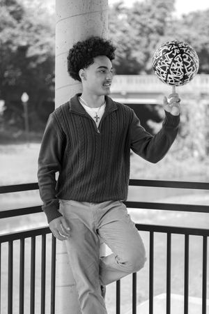 Black and white portrait of someone in a knit sweater balancing a decorative ball while posing by a railing outdoors.