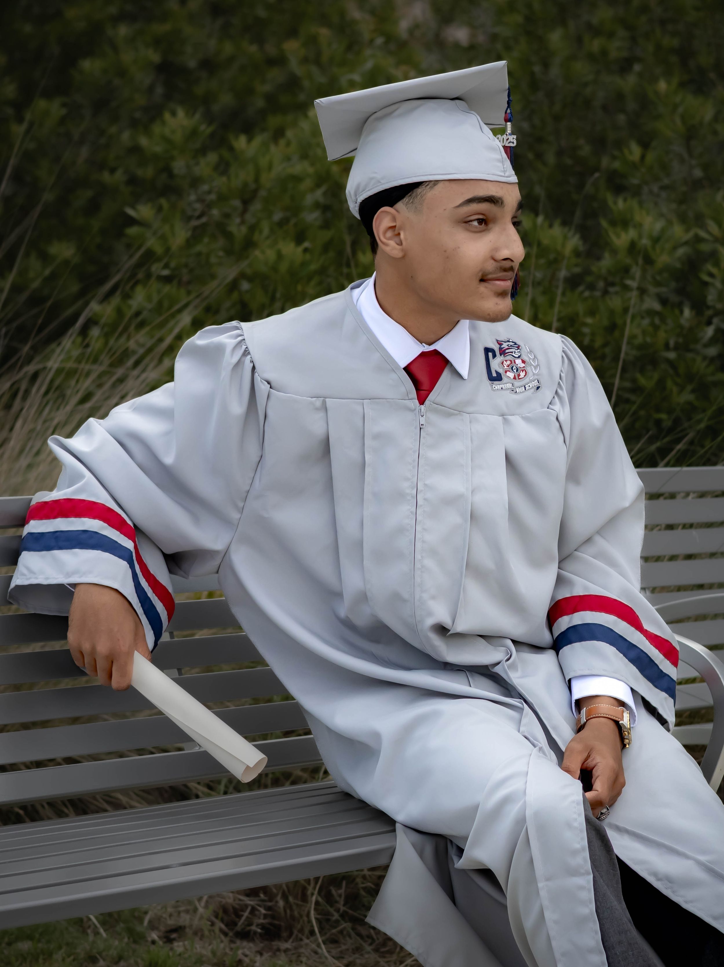 Graduate in white cap and gown with red tie sits casually on bench wearing patriotic striped sleeve details.