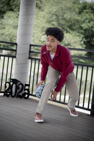 Person in burgundy sweater and khaki pants playfully poses on outdoor deck near white column.