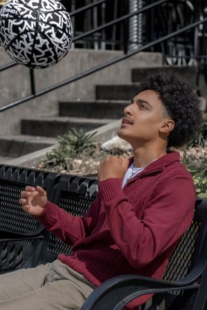 Person in burgundy zip-up jacket sits on bench looking up at decorative sphere with black letter patterns.