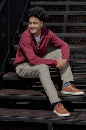 A young person in a burgundy sweater and khaki pants sits casually on wooden steps.