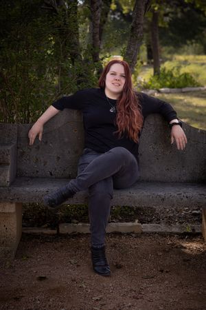 A person in dark clothing lounges casually on a stone wall outdoors with trees in the background.