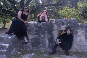 Three people in dark clothing pose together on a stone wall in an outdoor setting at dusk.