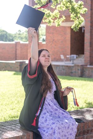 Young woman in graduation gown holding cap up triumphantly while sitting outdoors on a sunny day.