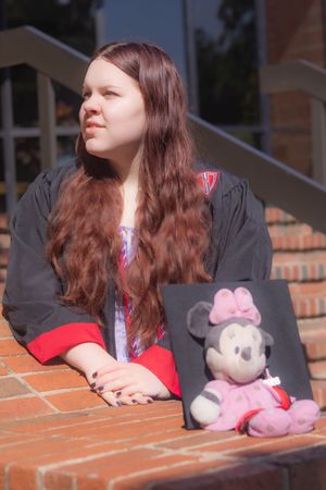 Young woman in graduation gown sits on brick steps holding Minnie Mouse plush toy outdoors.
