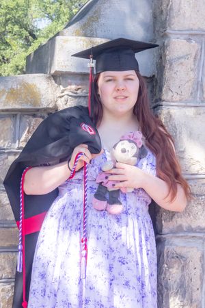 Young woman in graduation cap holding teddy bear and gown, posing by stone wall in floral dress.