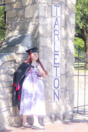 Female graduate in cap and gown poses by Tarleton State University stone entrance gate on sunny day.