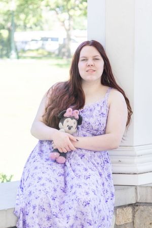 Young woman in lavender floral dress holding Minnie Mouse plush, leaning against white column outdoors.