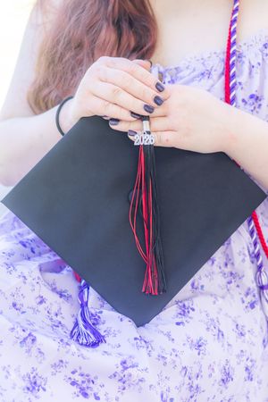 Young woman in floral dress holding a black 2020 graduation cap with red and blue tassels.