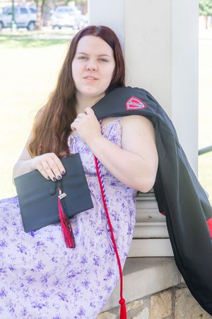 Young woman in floral dress holds graduation cap and gown with red tassel outdoors on campus.