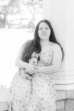 Black and white photo of a young woman in a floral dress holding a Minnie Mouse plush toy, leaning against a white column.
