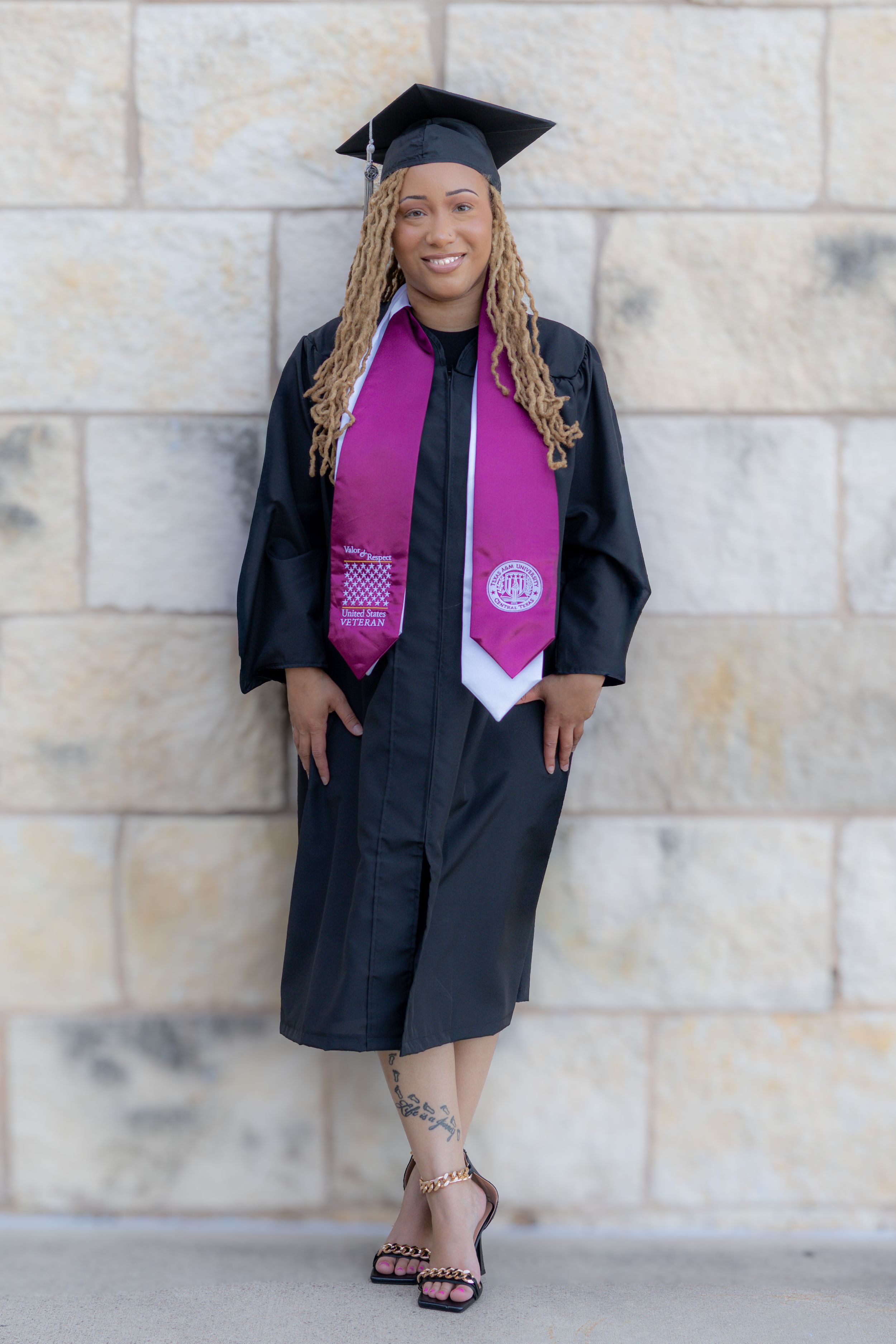 Graduate in black cap and gown with pink stole poses against stone wall in series of celebratory graduation photos.