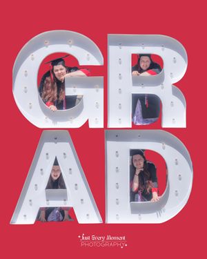 Graduation photo with four students in caps and gowns posing inside large illuminated GRAD letters on a red background.