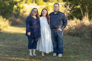 Three people smiling outdoors in a grassy field, two women and a man posing together for a family portrait.