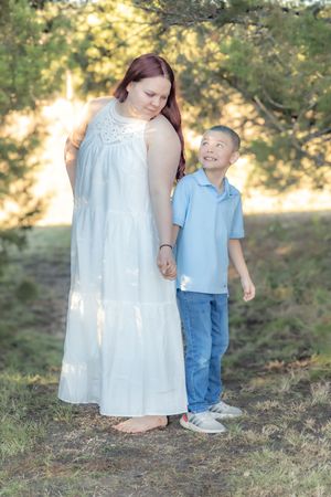 A child in blue shirt and jeans plays outdoors with someone wearing a white dress in a sunlit wooded setting.
