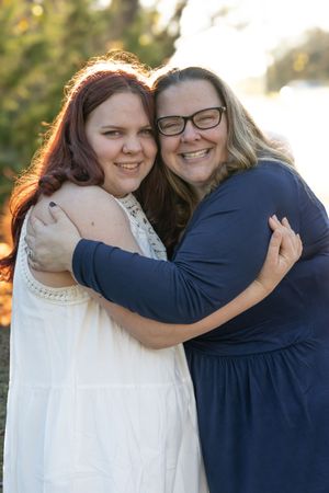 Two people embrace warmly while smiling outdoors in golden sunlight, one wearing white and one in navy blue.