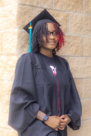 A smiling graduate in black cap and gown with teal tassel and red-highlighted locs poses outdoors.
