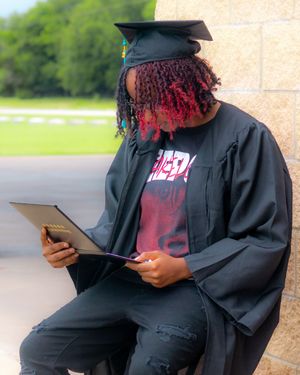 Graduate in black cap and gown sits reading diploma, wearing ripped jeans and red graphic tee outdoors.