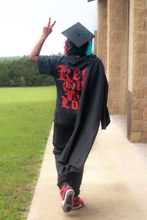 Graduate in black cap and gown with red graphic tee flashing peace sign while walking on campus sidewalk.