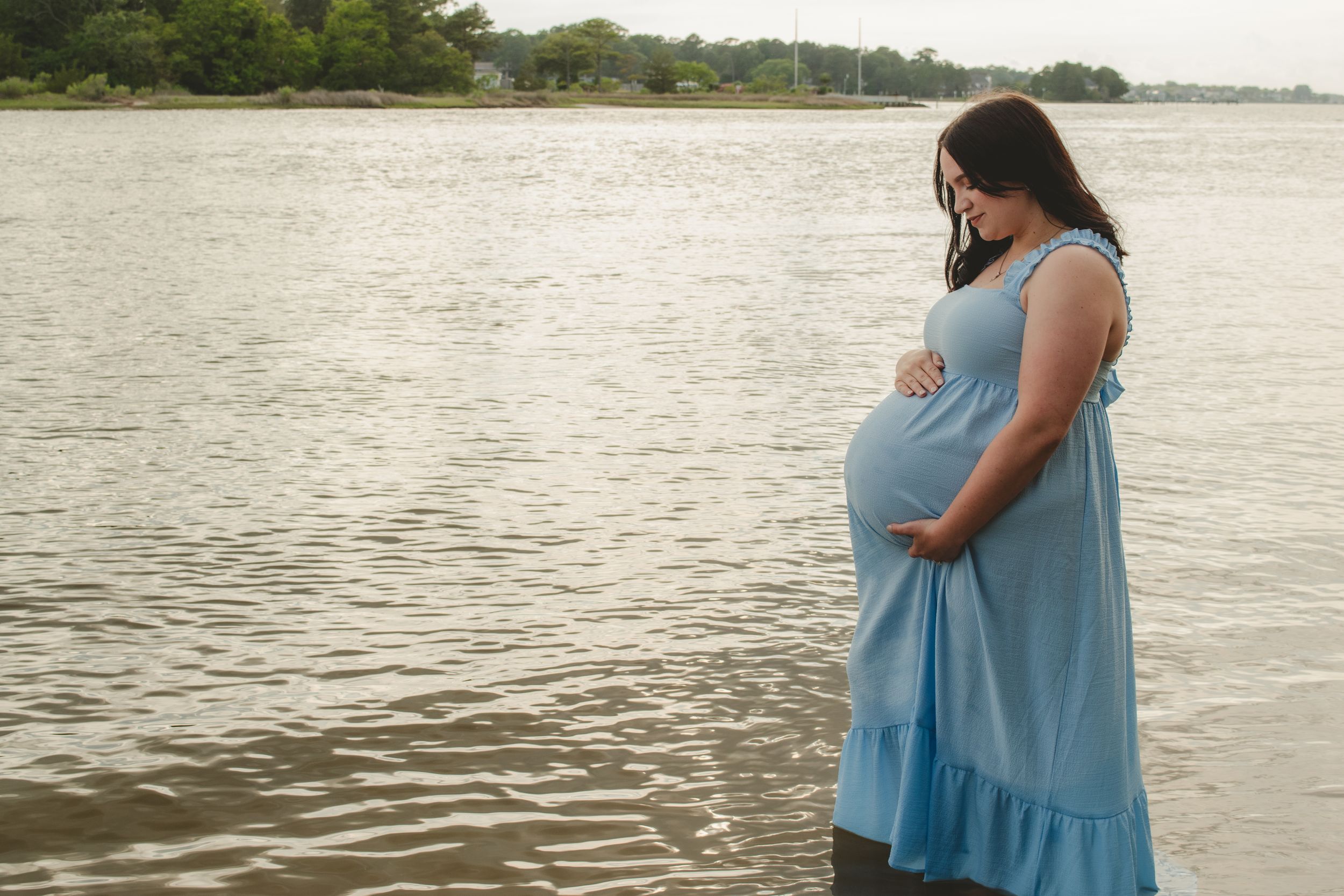 Pregnant woman in blue dress stands reflectively by calm waterside during golden hour.