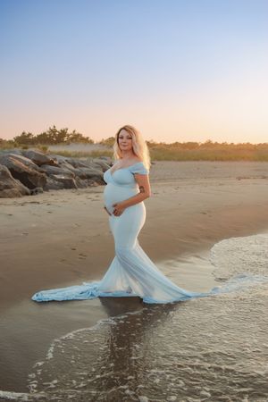 Pregnant woman in flowing light blue gown poses elegantly on beach at sunset with waves lapping at her feet.