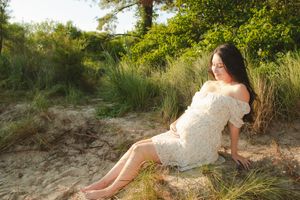 Someone in a white lace dress sits gracefully on sandy ground near green foliage during golden hour lighting.