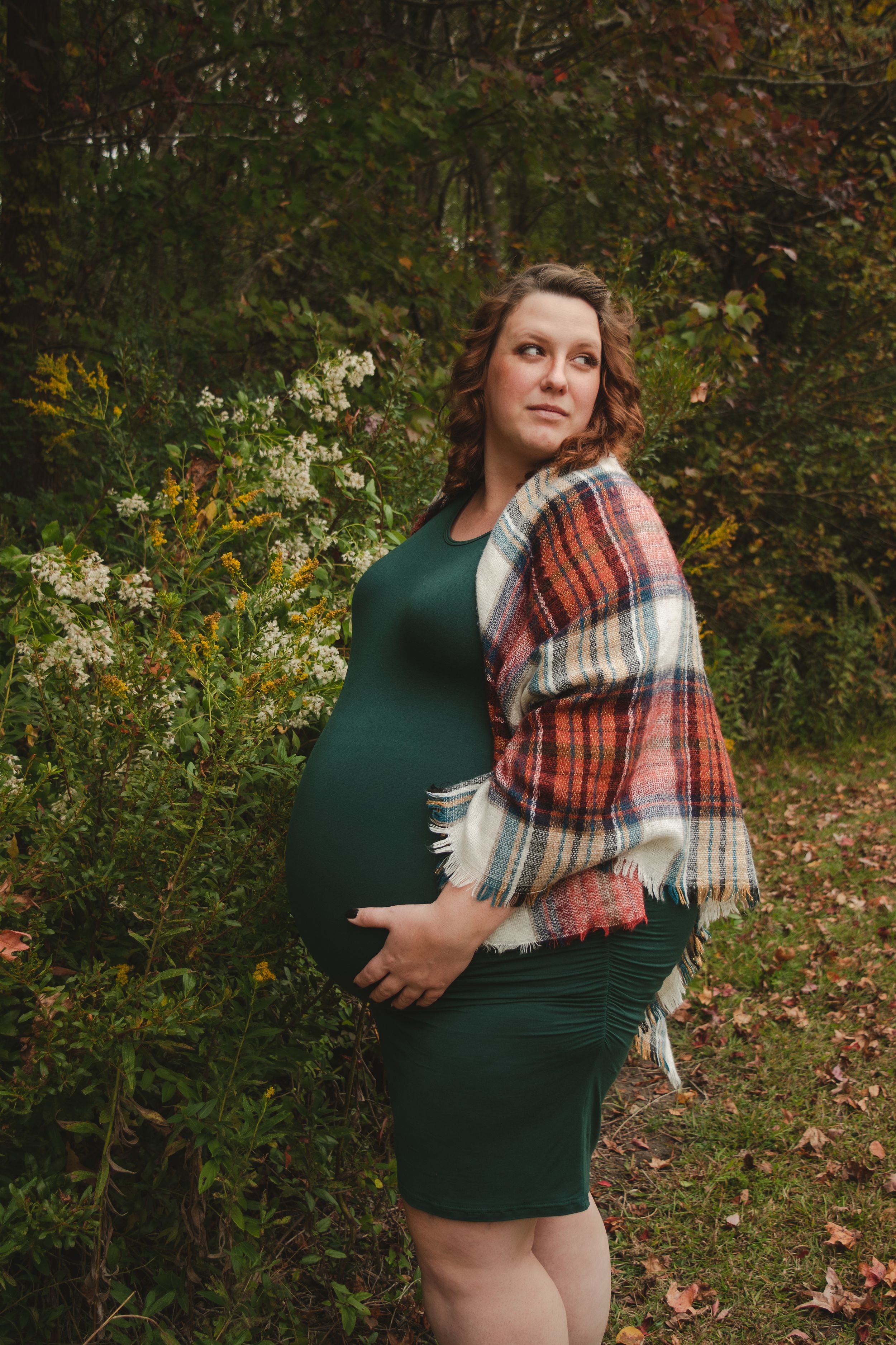 Maternity photo session in autumn forest with woman in green dress holding plaid blanket.