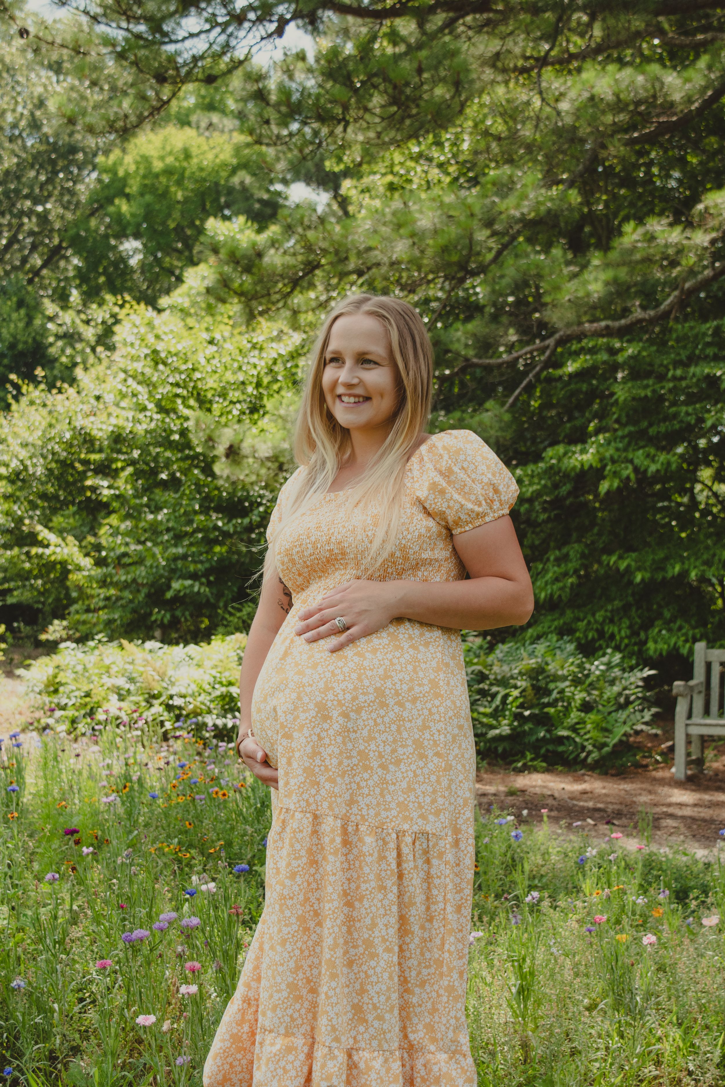 Expectant woman in a flowing yellow floral dress stands in a garden with wildflowers and greenery on a sunny day.