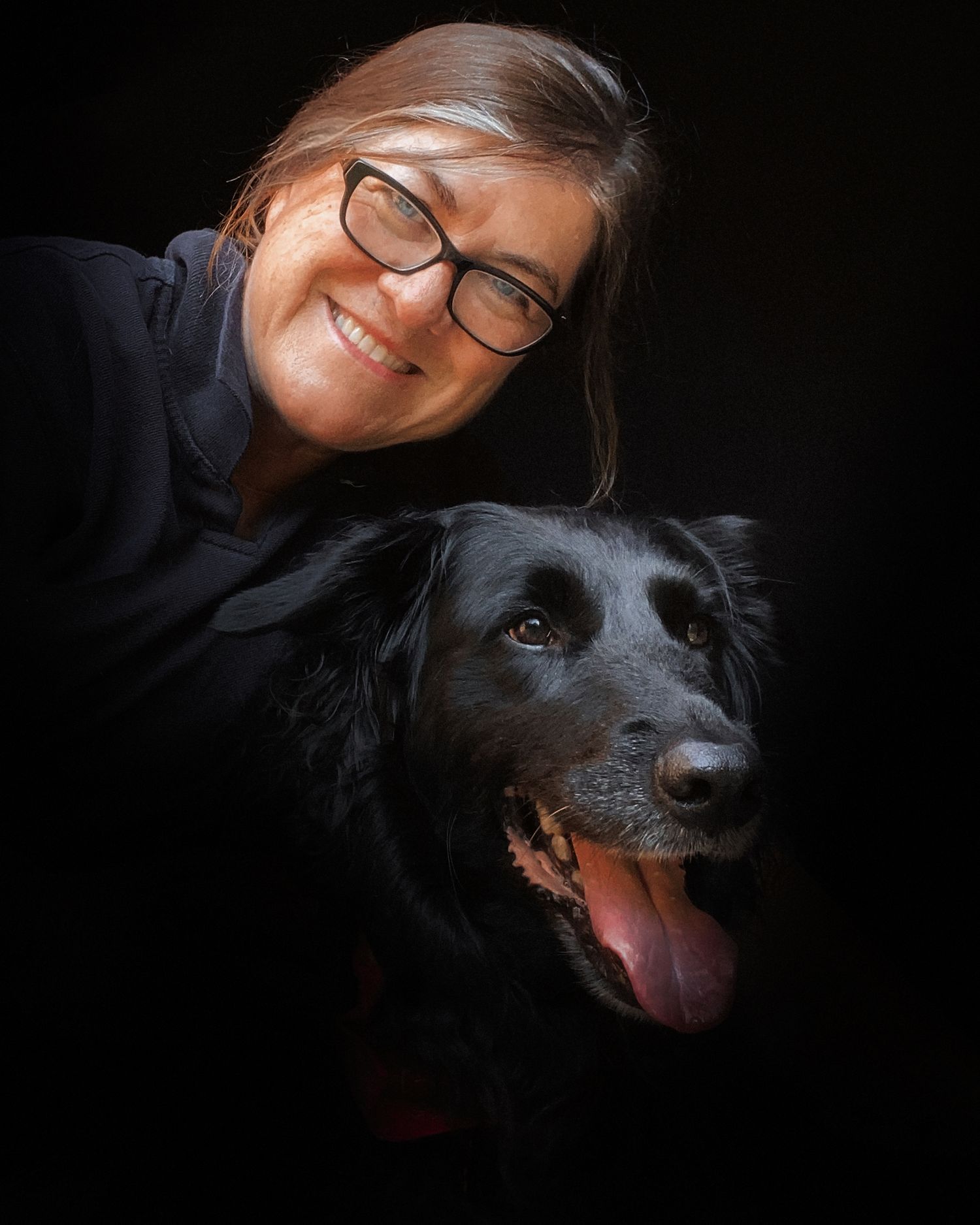 A joyful portrait of someone with glasses posing with their happy black dog against a dark background.