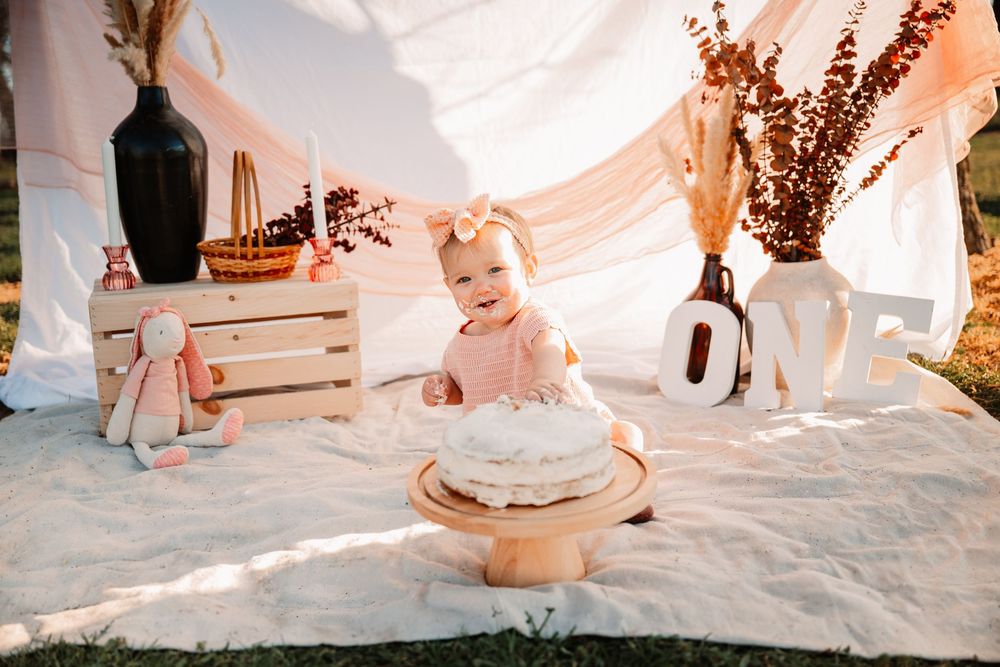 First birthday milestone photo of baby with cake and ‘ONE’ sign in Dallas–Fort Worth with florals, a rabbit and a white sheet hanging behind
