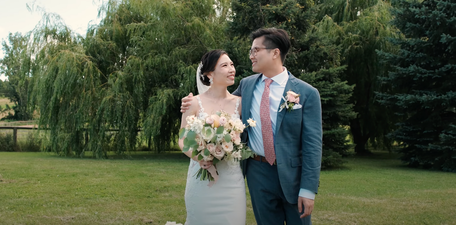Newlyweds pose together outdoors with a summer garden backdrop of willow trees and lush greenery.