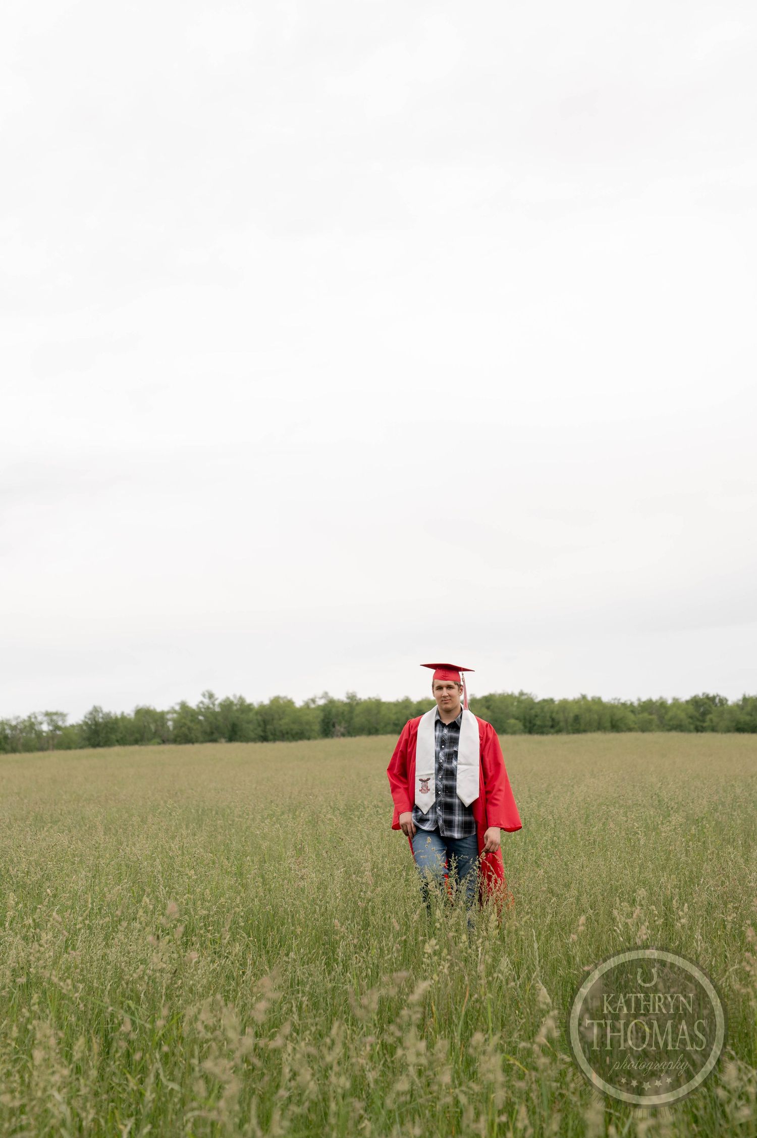 Graduate walks through a green prairie field wearing academic regalia with trees visible along the horizon.