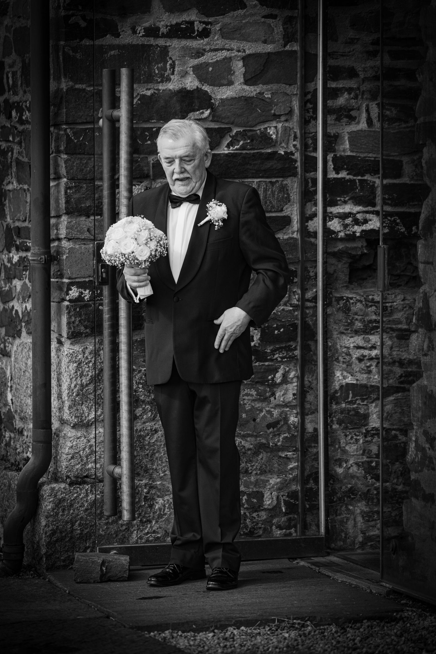 Elegant elderly man in tuxedo holding bridal bouquet outside stone church wall.