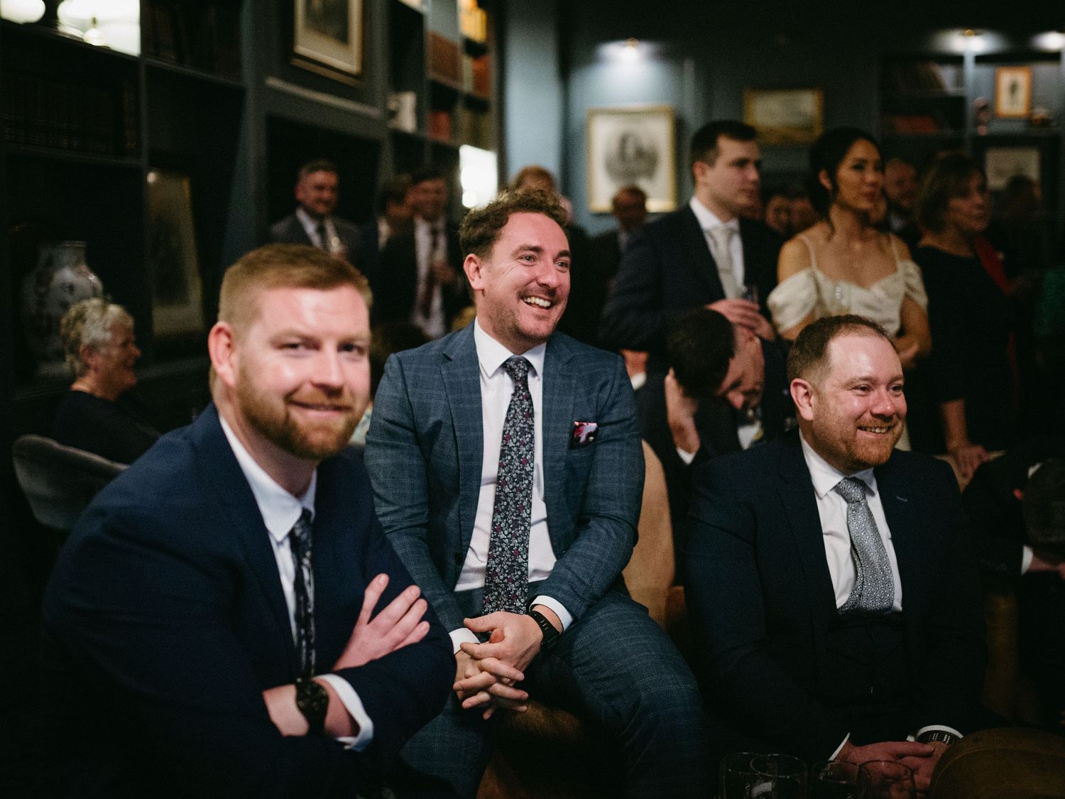 A group of men in suits share laughter and conversation at a formal indoor gathering.