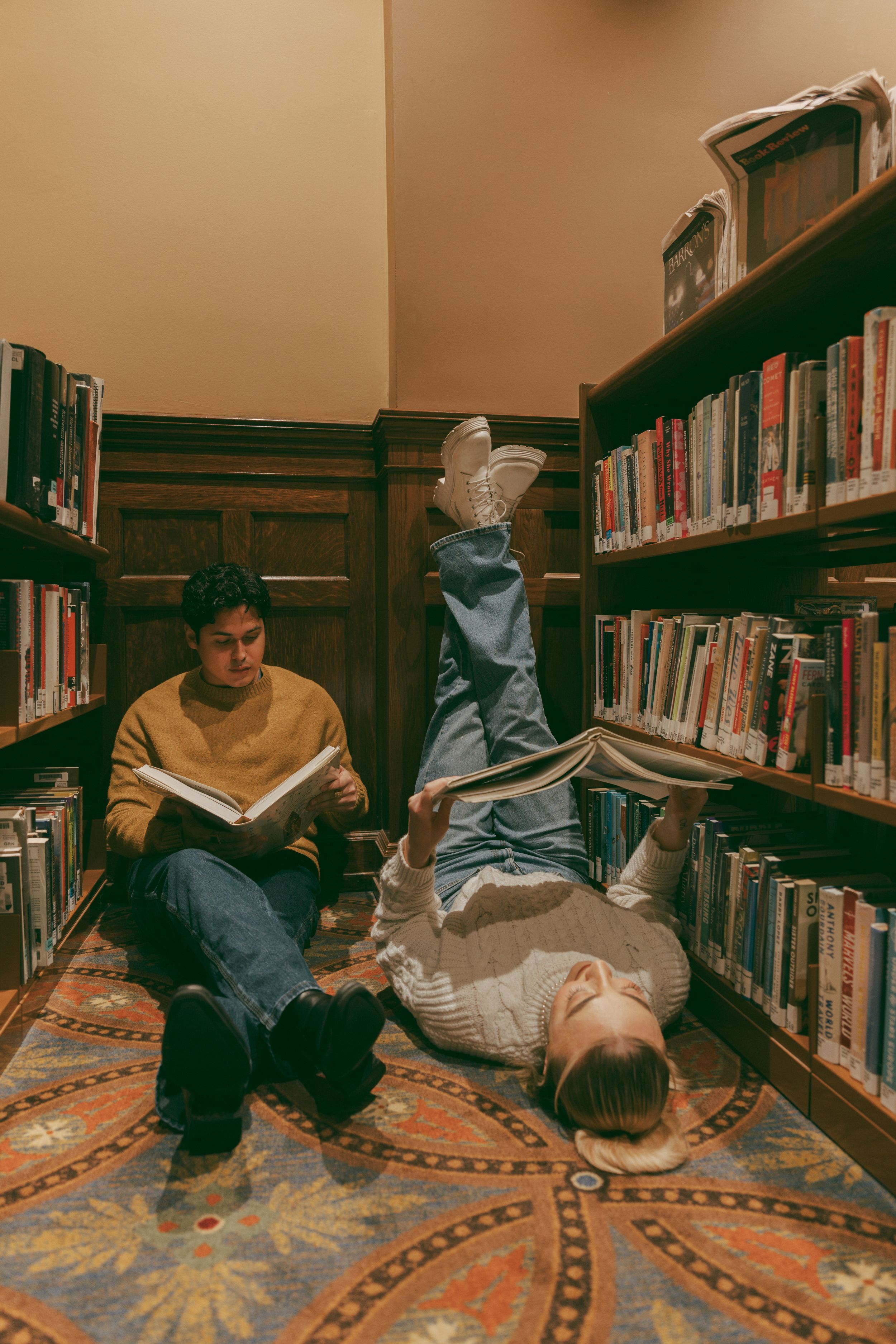 Two young people relaxing with books in a library corner with dark wood shelves and a decorative patterned carpet.