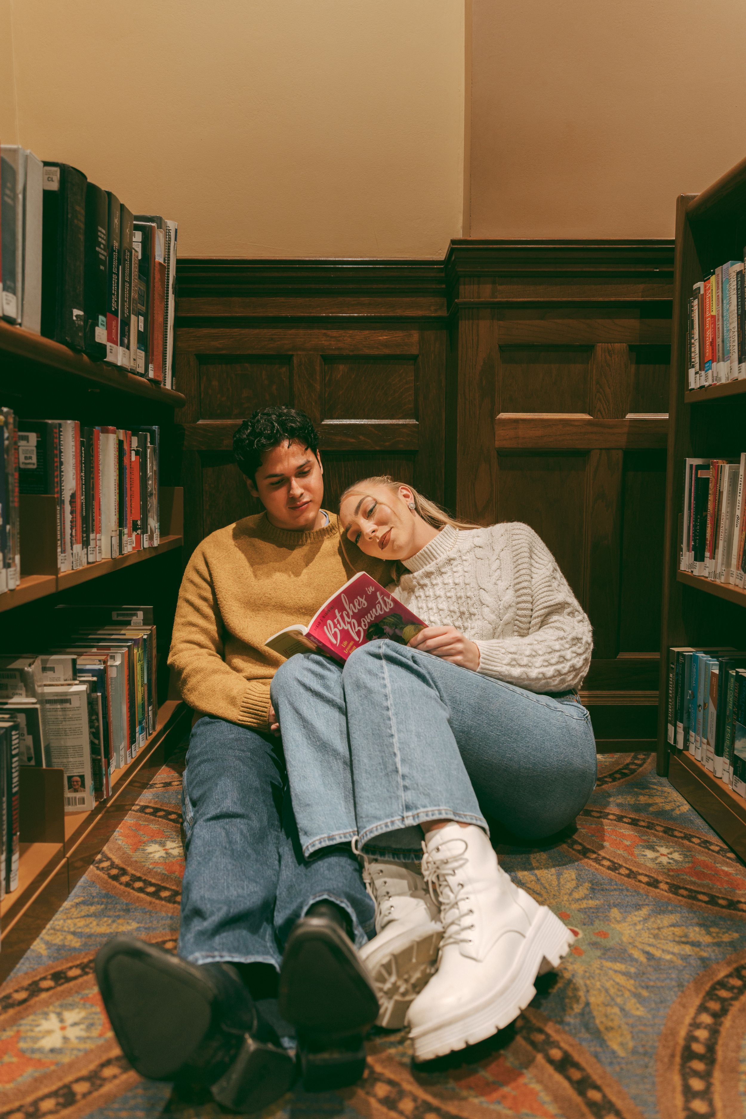 Two people sitting on a carpeted library floor between bookshelves, one resting on the other's shoulder while holding a book.