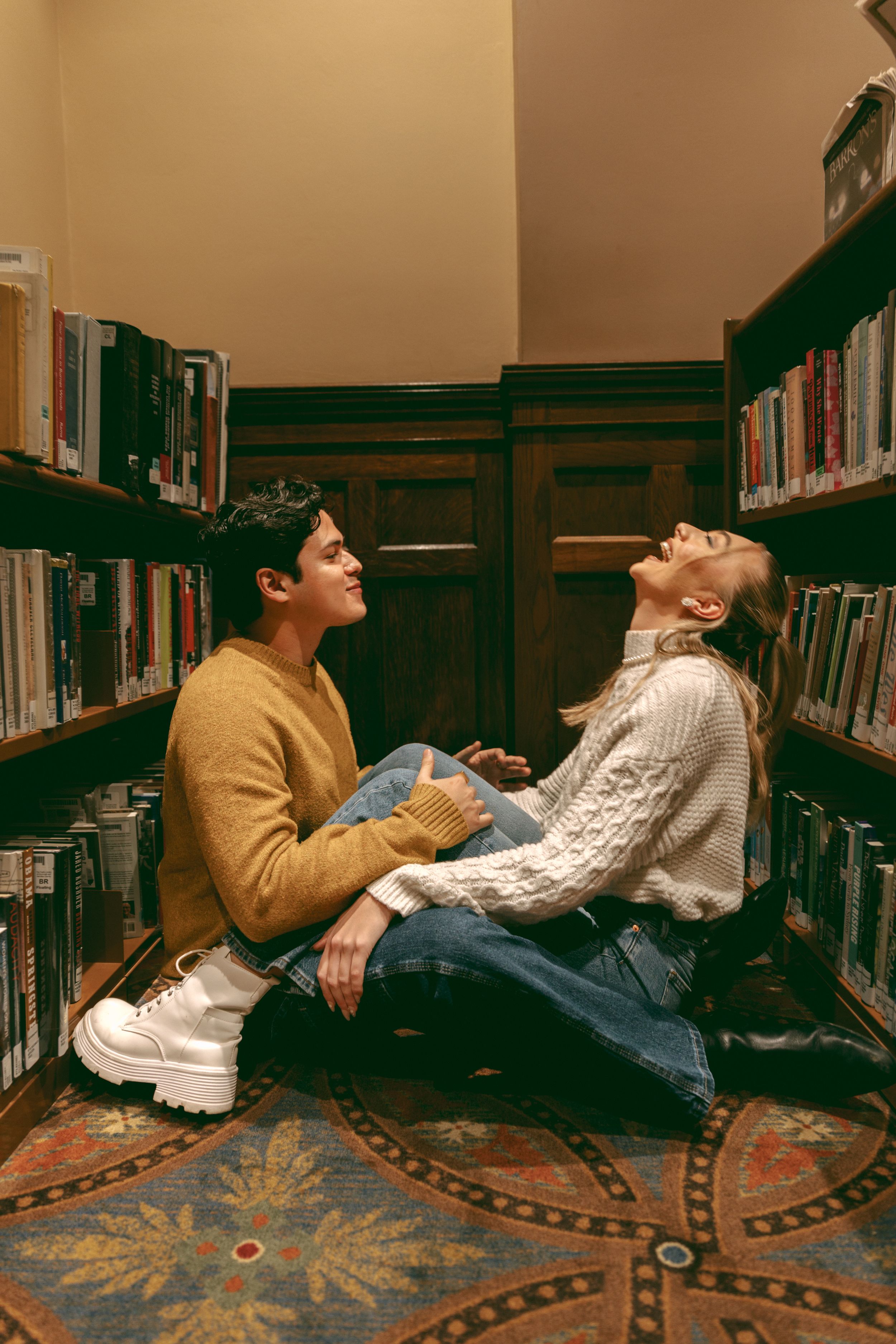 Two people laughing together while sitting on the floor of a library between bookshelves with a decorative carpet beneath them.