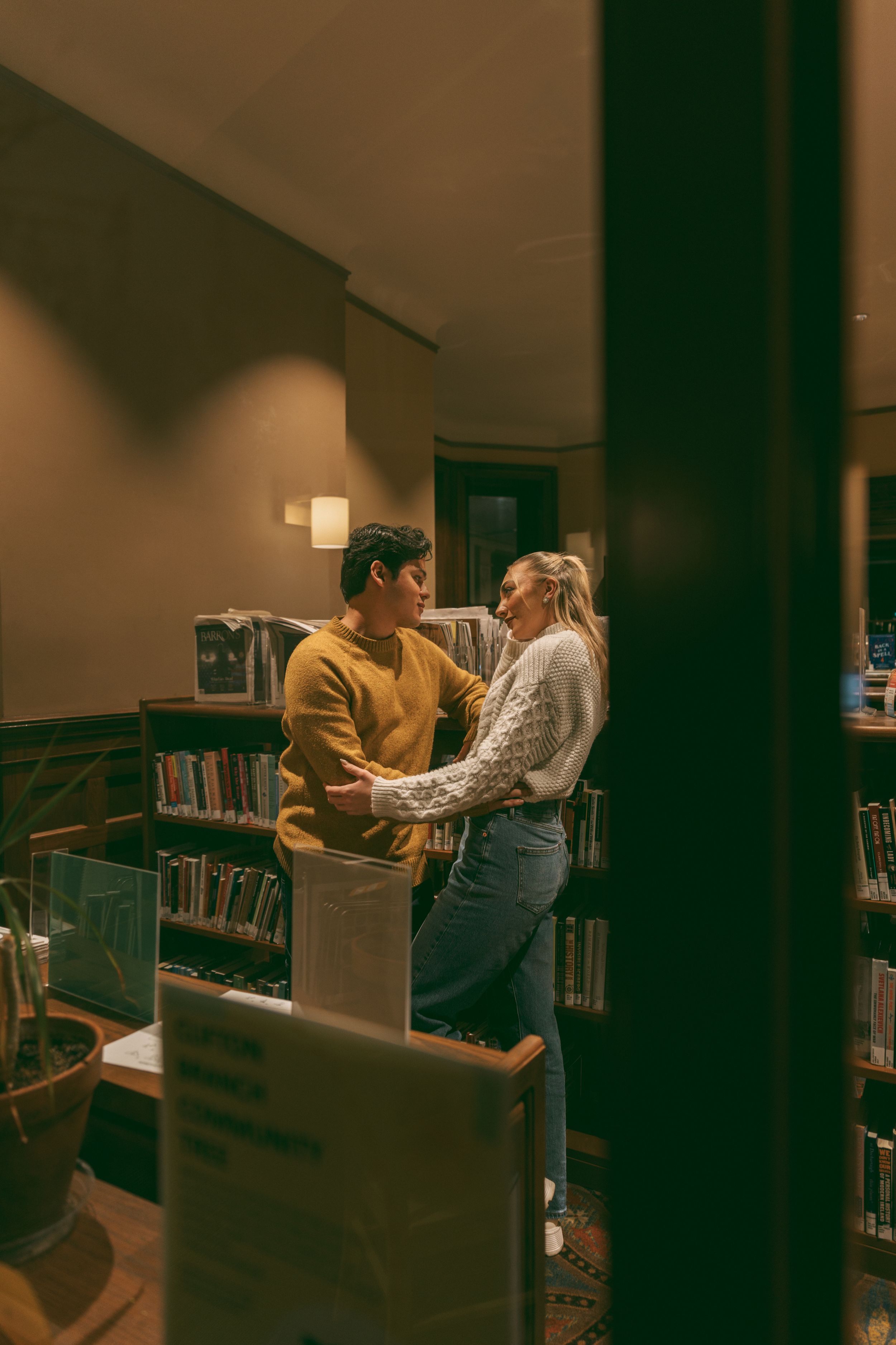 A romantic couple embraces in a dimly lit library setting surrounded by bookshelves.