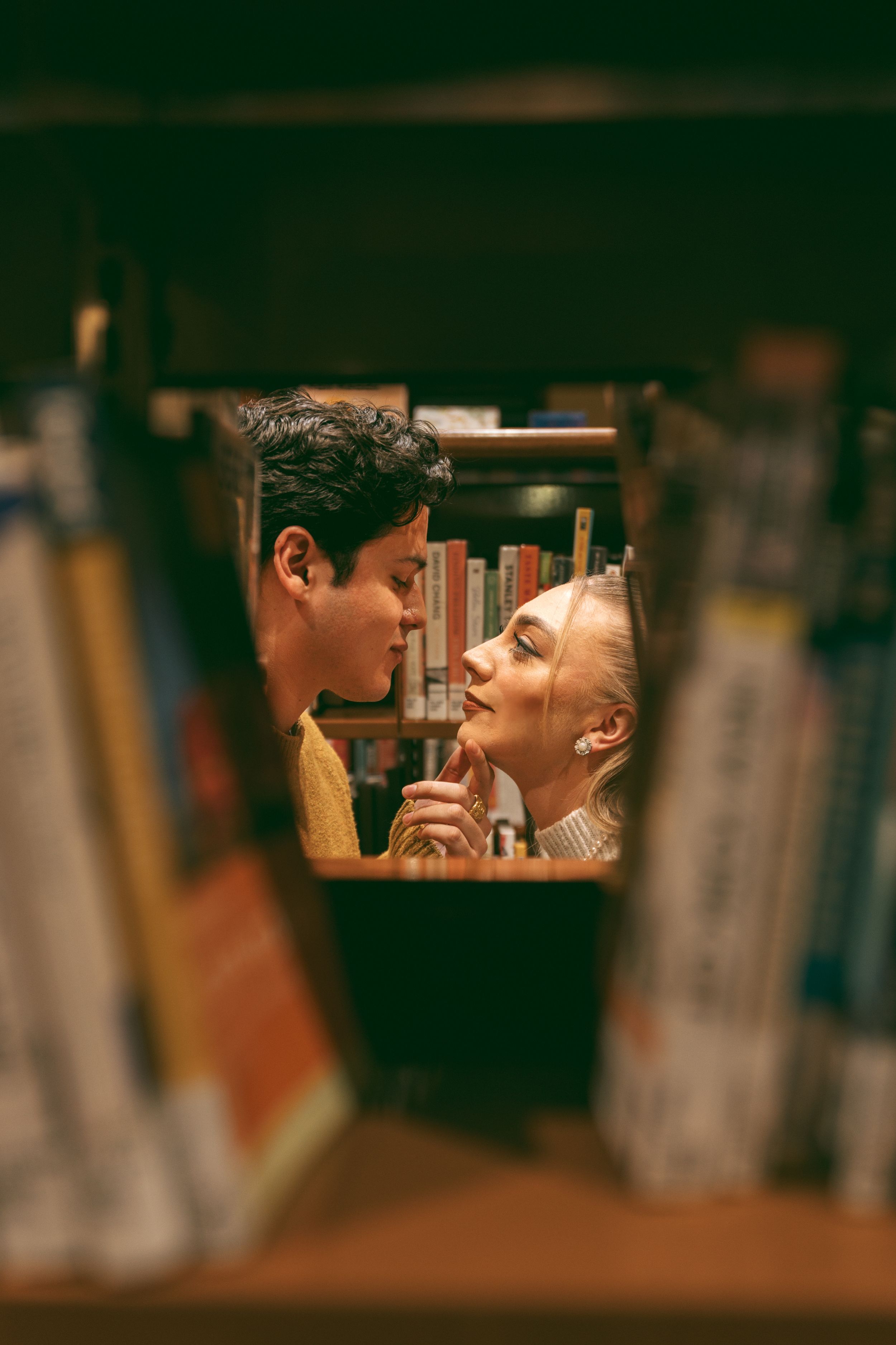 A couple shares a romantic moment between library bookshelves in warm lighting.