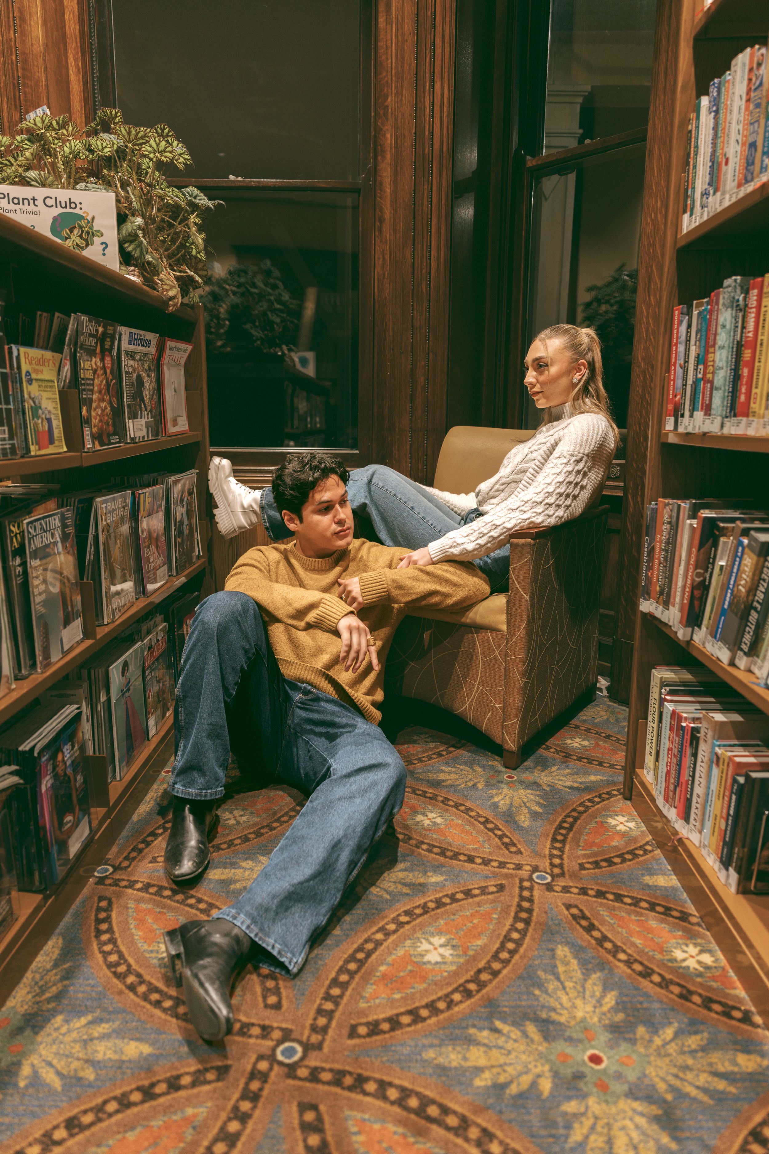 A couple lounging playfully between library bookshelves on a patterned carpet floor.