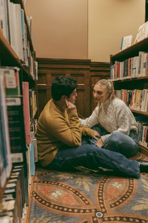 Couple sharing a tender moment while sitting on library floor between wooden bookshelves.