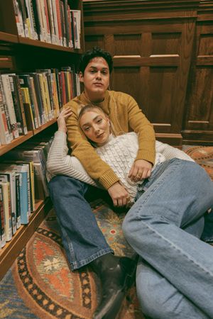 Two people relaxing intimately among library shelves on an ornate rug.