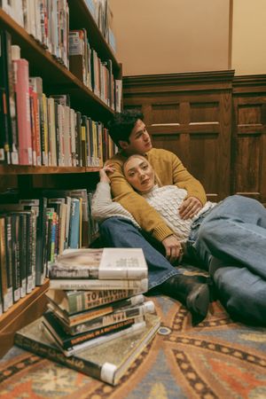 Couple cuddling on library floor surrounded by wooden shelves and scattered books.