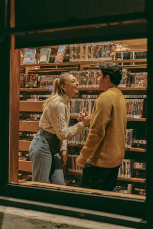 Two people laughing together between library shelves filled with books and media.
