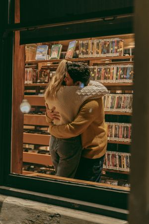 Couple in warm sweaters embracing between library shelves.